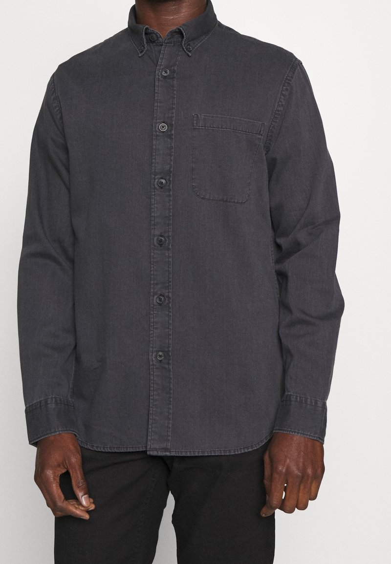 Homme portant une chemise à manches longues gris foncé boutonnée avec une poche poitrine avant et un pantalon noir sur un fond blanc uni.