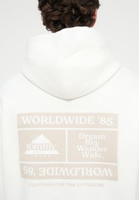 Λευκή ζακέτα με μεγάλη μπεζ γραφιστική στο πίσω μέρος που περιλαμβάνει κείμενο και λογότυπο βουνού. Το ύφασμα φαίνεται μαλακό με βουρτσιστή υφή.