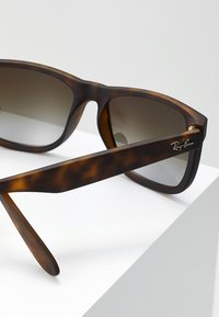 Ochelari de soare cu carapace de broască țestoasă, cu lentile maro cu gradient. Caracteristicile includ un cadru gros, o textură netedă și un logo „Ray-Ban” auriu pe braț.