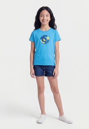 Fată zâmbitoare cu păr lung și negru, îmbrăcată într-un tricou albastru cu imprimeu, pantaloni scurți din denim închis și adidași albi, stând în fața unui fundal simplu.