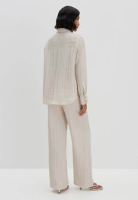 Camicia leggera beige, con texture, colletto button-down, maniche lunghe e pieghe sul retro, abbinata a pantaloni larghi coordinati.