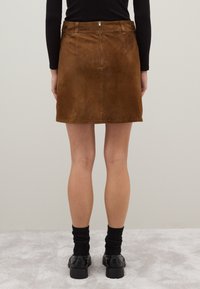Mujer vestida con una mini falda de ante marrón con cremallera trasera, calcetines negros, zapatos negros y una blusa negra de manga larga, de pie sobre una alfombra clara.