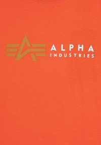 Narančasta majica s kratkim rukavima koja ima zlatni logo s krilima i bijeli natpis "ALPHA INDUSTRIES". Glatka tkanina s okruglim izrezom.