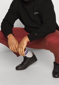 Svart huvtröja med logotyp, vinröda byxor, svarta sneakers och vita strumpor med blå ränder. Fokus på händer i en hukad position.