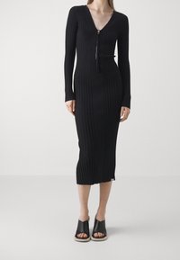 Robe en maille côtelée noire avec un décolleté en V et des manches longues, dotée d'une fermeture éclair frontale. La robe descend en dessous du genou et présente une coupe ajustée.