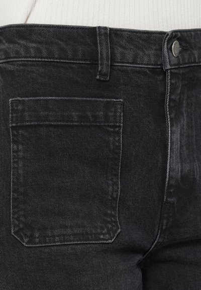 Aizmugures skats uz tumši melnām džinsa biksēm ar kabatu, jostas cilpām un metāla pogas aizdari, valkājot baltu ribota auduma topu.