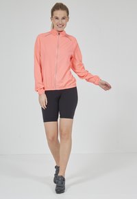 Ljus korallfärgad zip-up sportjacka med hög krage och elastiska muddar, i kombination med svarta cykelshorts och svarta cykelskor.