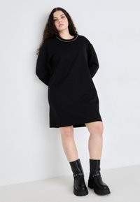 Čierne oversize šaty s okrúhlym výstrihom, so striebornými detailami. Kombinované s čiernymi robustnými čižmami na podpätku. Hladká štruktúra látky.
