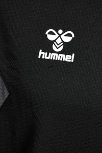 Maglietta atletica nera realizzata in tessuto testurizzato, con un logo bianco che presenta un design di ape e il nome del marchio "hummel" al centro. Accentuata da dettagli grigi.