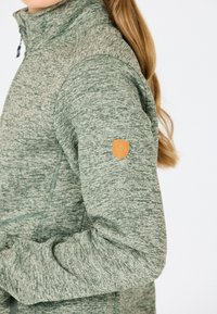 Kvinna i ljusgrön melerad zip-up jacka med en tan läderpatch på övre ärmen, håret uppsatt i en lös hästsvans.