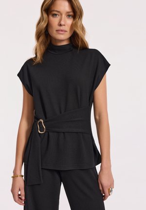 Kobieta ubrana w czarną bezrękawnikową bluzkę o wysokim dekolcie, z szerokim pasa i złotą klamrą, w zestawieniu z czarnymi spodniami na jednolitym tle.