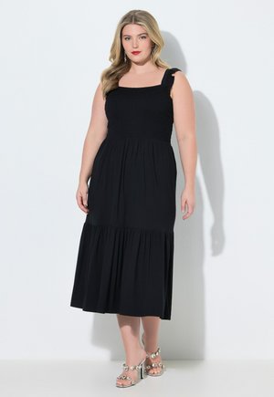 Kvinde med bølget blondt hår iført en sort ærmeløs midi-kjole med flæser på stropperne og sølvfarvede højhælede sandaler med juvelbesætning.