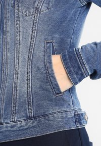 Chaqueta de mezclilla en un lavado azul medio con costuras en contraste, que incluye un bolsillo frontal, puños ajustados y un largo ligeramente recortado.