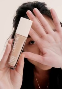 Lancôme foundationflaske med klar glassbase og hvit hette, som viser en lys beige væske inni; en hånd blokkerer delvis utsikten.