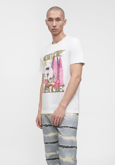 Άνδρας που φοράει λευκό γραφικό t-shirt με αετό, ουρανοξύστες και αστέρια, σε συνδυασμό με γκρι και κίτρινες παντελόνες tie-dye σε απλό φόντο.