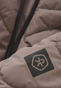 Chaqueta acolchada de color marrón claro con una textura suave. Presenta un parche de logo negro y un cierre de cremallera en el lateral.