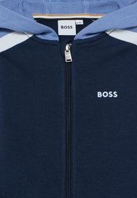 Μπλε ναυτικό φούτερ με φερμουάρ, διαθέτει κουκούλα σε γαλάζιο χρώμ α, ριμπ υφή και ένα μικρό λευκό λογότυπο "BOSS" στο αριστερό μέρος του στήθους.