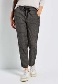 Vrouw in grijze geruite joggers en witte sneakers, staand met één hand in haar zak tegen een effen witte achtergrond.