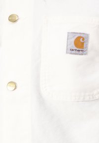 Camisa de mezclilla blanca con un bolsillo en el pecho que tiene una etiqueta gris y un logo naranja, terminada con botones dorados para dar detalle.