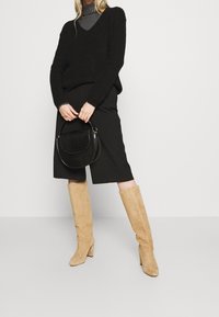 Schwarzer gestrickter V-Ausschnitt Pullover, grauer Rollkragenpullover darunter, schwarzer Midirock, sandfarbene Wildleder-Stiefel bis zum Knie und eine schwarze Schultertasche mit Krokodilprägung.