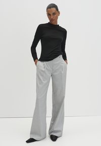 Frau in einem schwarzen Langarm-Oberteil, hellgrauen, weit geschnittenen Hosen und schwarzen spitzen Schuhen, die mit den Händen in den Taschen vor einem weißen Hintergrund steht.