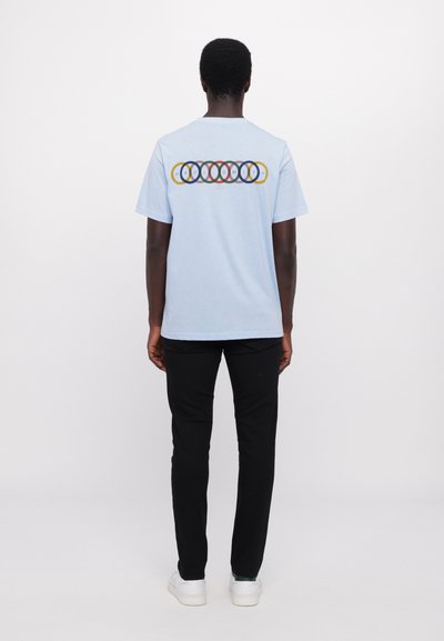 Lyseblå kortærmet t-shirt med et multikolor cirkulært logo og "PAUL SMITH" tekst på ryggen. Båret med sorte bukser og hvide sko.