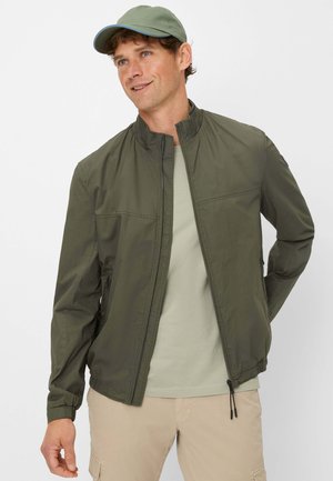 Olive green lightweight jacket with a standing collar, zip closure, and side pockets. Paired with a light green t-shirt and beige pants.