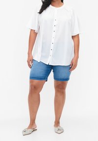 Femme portant un chemisier blanc ample à boutons, un short en jean bleu longueur genou, et des chaussures à enfiler pointues avec un détail nœud.