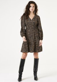 Vestido negro con patrón floral dorado, escote en V, mangas largas abullonadas y cintura fruncida. Llevado con botas negras hasta la rodilla.