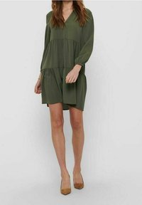 Olivengrünes Kleid mit langen Ärmeln, V-Ausschnitt, schichtweise Design und weichem Stoff. Kombiniert mit beige-farbenen, spitz zulaufenden Absatzschuhen.