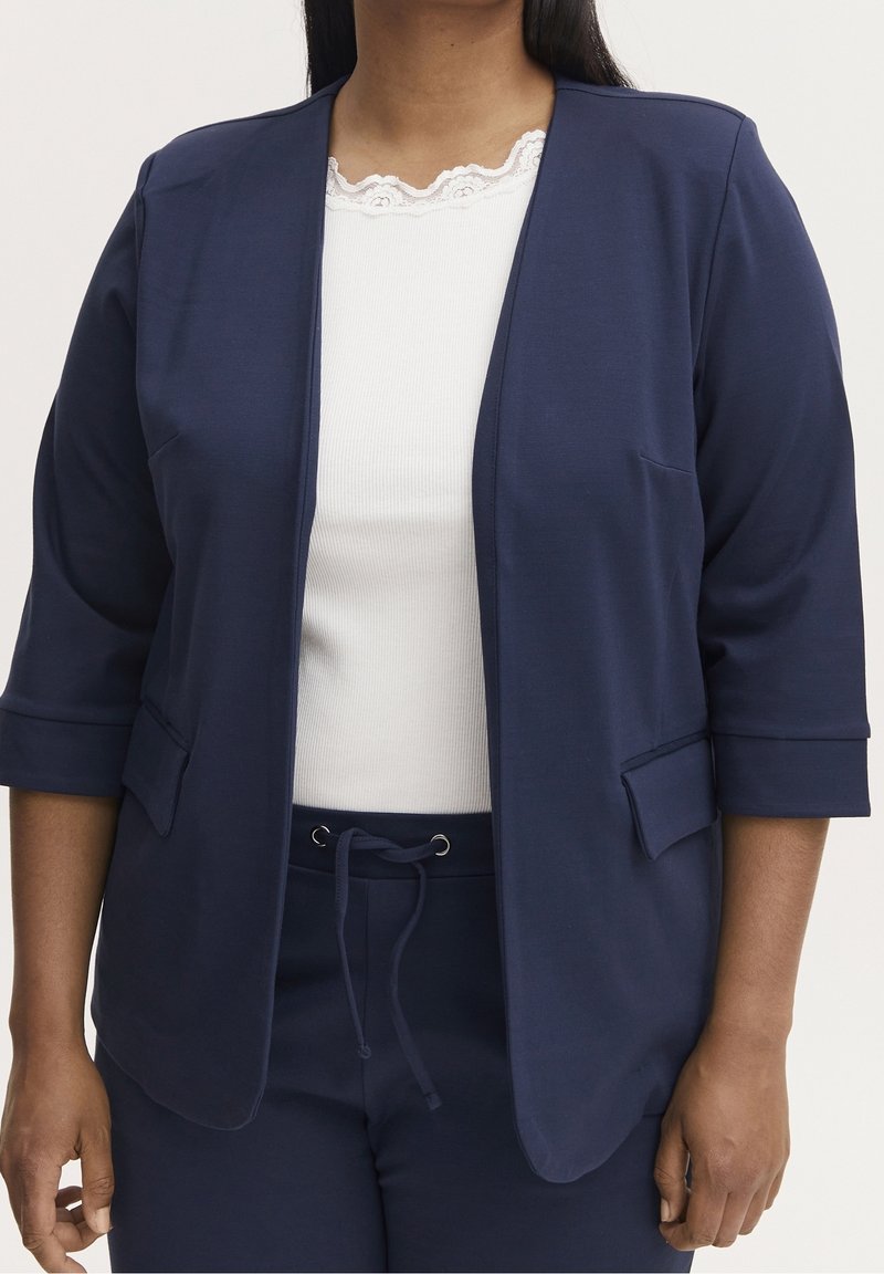 Femme portant un blazer bleu marine ouvert sur le devant avec des manches trois-quarts, un top blanc bordé de dentelle, et un pantalon bleu marine à cordon.