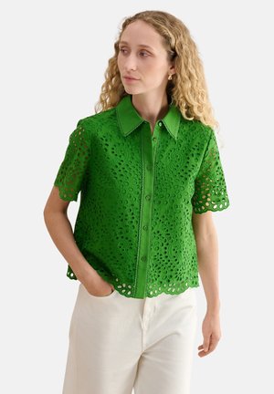 Mujer con cabello rubio rizado que lleva una blusa verde brillante de manga corta con ojales y pantalones blancos, mirando hacia un lado con una mano en el bolsillo.