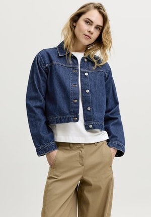 Femme aux cheveux blonds portant une veste en jean courte bleu foncé, un t-shirt blanc et un pantalon beige, debout les mains dans les poches.