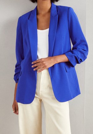 Person iført en knaldbla blazer med opkørt ærmer, en hvid top og lysebeige bukser, stående mod en ensfarvet baggrund.