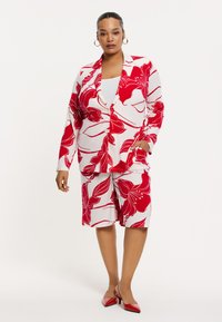 Bloemendessin outfit met een rood-witte blazer met grote bloemen, gecombineerd met bijpassende shorts. Glanzende rode hakken maken de look af.