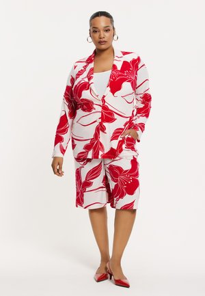 Blumenmuster-Outfit mit einem rot-weißen Blazer mit großen Blumen, kombiniert mit passenden Shorts. Glänzende rote High Heels runden den Look ab.