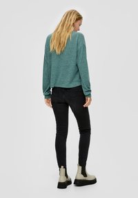 Turkusowy, długi sweter z krótkim krojem i luźnym fasonem, zestawiony z czarnymi skinny jeansami i beżowymi botkami za kostkę na grubej podeszwie. Gładkie tło.