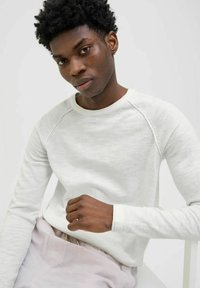 Camicia bianca a maniche lunghe con scollatura tonda e maniche raglan, realizzata in un morbido tessuto testurizzato. Il modello posa su uno sfondo neutro.