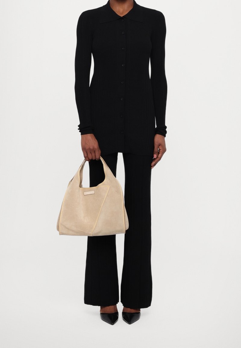 Personne portant un haut noir à manches longues boutonné et un pantalon, tenant un grand sac à main beige doux avec des poignées courtes.