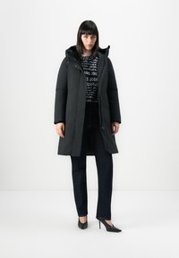 Vrouw staat in een lange zwarte pufferjas, een zwart-wit gestreepte trui, donkere jeans en zwarte pointed high heels tegen een witte achtergrond.