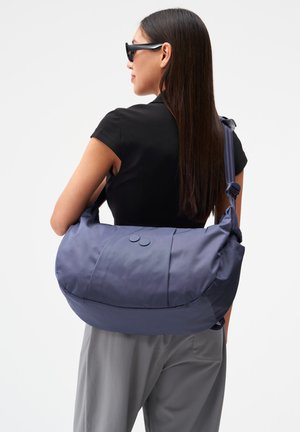 Frau mit langen dunklen Haaren, die Sonnenbrillen trägt, schwarzes Oberteil, graue Hose, trägt eine große dunkelblaue Umhängetasche mit zwei Knöpfen auf der Rückseite.