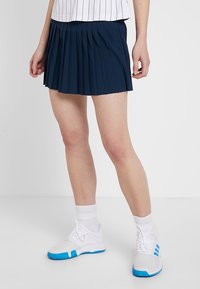 Marinblå plisserad kjol av lätt tyg. Har en figursydd midja och slutar mitt på låret. Bärs med en vit topp med ränder och träningsskor.