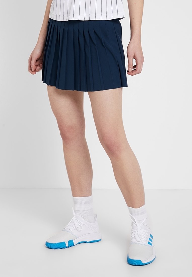 Marinblå plisserad kjol av lätt tyg. Har en figursydd midja och slutar mitt på låret. Bärs med en vit topp med ränder och träningsskor.