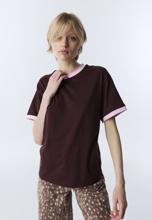 Blond osoba nosząca ciemnobrązowy T-shirt z różowym obszyciem i brązowe spodnie z białymi plamami, stojąca na jednolitym jasnym tle.