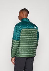 Padded jacket s modro-zelenou horní částí a zeleným spodním dílem, s horizontálním šitím, vyrobená z lesklého, lehkého materiálu.