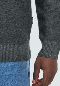 Mørk grå strikkegenser med ribbestrikkede mansjetter og søm, med en strukturert veving. Merket på siden sier "BLEND." Bærer over blå denimjeans.