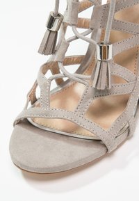 Sandale en daim gris clair avec bout ouvert, brides croisées, lacets à pompons avec embouts en métal argenté, et semelle intérieure en cuir beige.