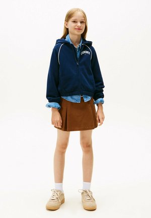 Felpa navale con zip e accenti bianchi, gonna plissettata marrone, camicia colletto azzurro chiaro, sneakers beige con suola testurizzata e calzini bianchi.