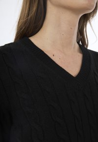 Maglione nero a maglia con scollo a V, texture a trecce e bordi a costine. La ripresa ravvicinata mostra il dettaglio complesso del motivo a trecce.