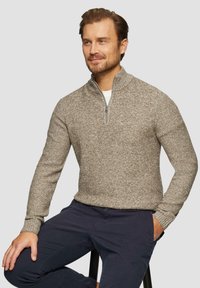 Ein Mann sitzt auf einem Hocker und trägt einen braunen Halbzip-Pullover über einem weißen Hemd und einer blauen Hose, mit einer Hand in der Tasche und einem leichten Lächeln.